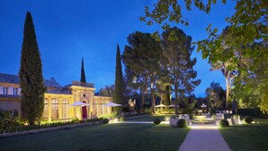 Un jardín bien iluminado con un sendero que lleva a un edificio de fachada tradicional y un árbol alto y esbelto en primer plano.