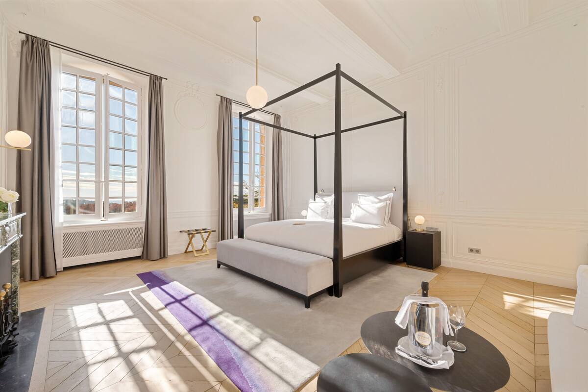Ein modernes Schlafzimmer mit einem Himmelbett, einem großen Fenster mit Vorhängen, einem Nachttisch mit Lampe und einem kleinen Beistelltisch.
