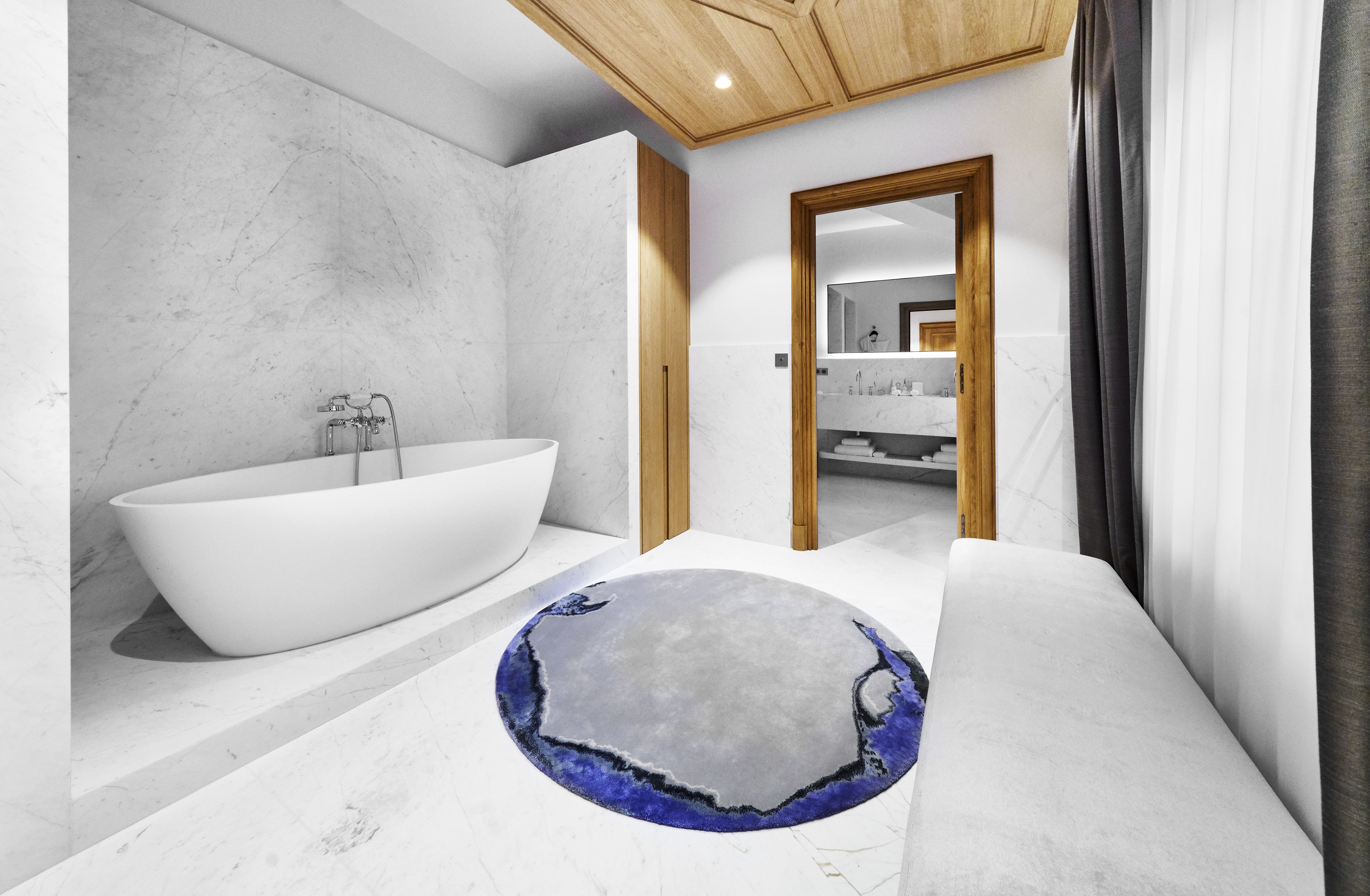 A modern bathroom with a freestanding bathtub, a round rug with a unique design, and a wooden ceiling.