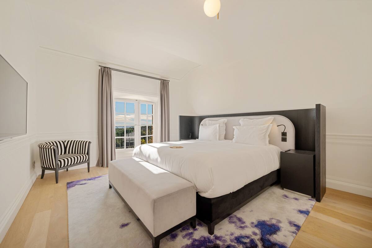 Ein Schlafzimmer mit einem großen Bett, einem Sessel, einem Nachttisch und Blick ins Freie.