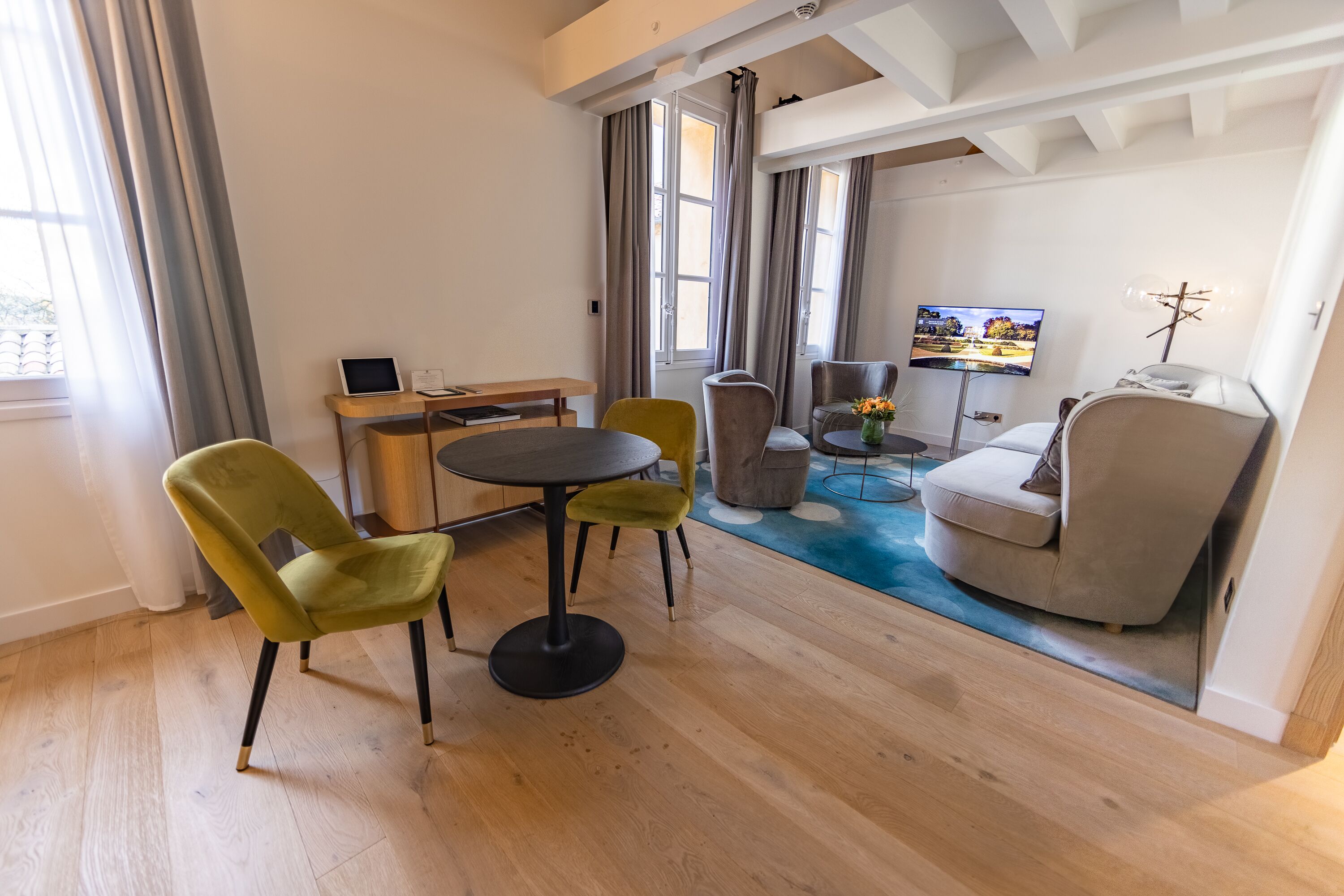A modern living room with a wooden floor, a round table, two green chairs, a grey sofa, and a TV mounted on the wall.