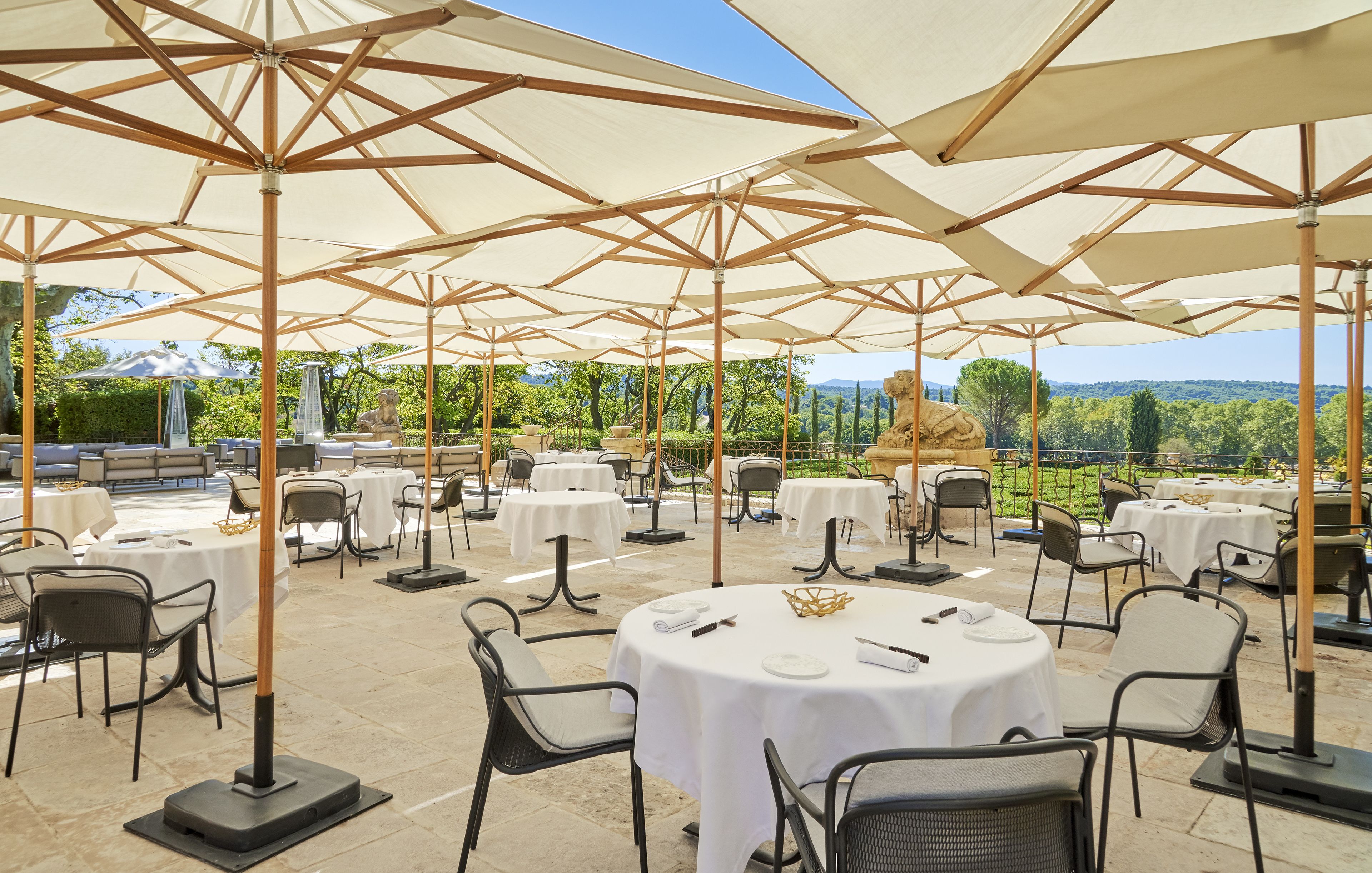Een terras met ronde tafels en stoelen, beschaduwd door grote parasols, met een achtergrond van bomen en heuvels.