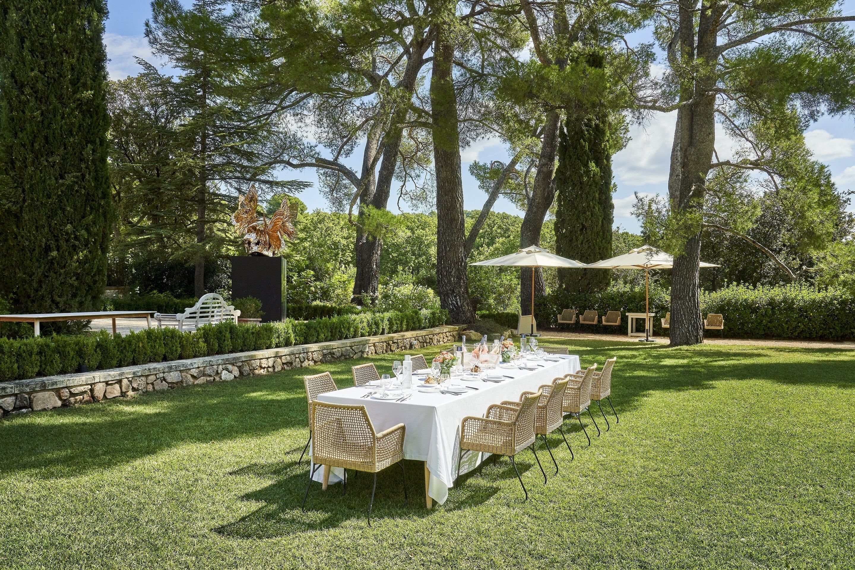 An outdoor dining area with a long table set for a meal, surrounded by chairs and umbrellas, with trees and a stone wall in the background.