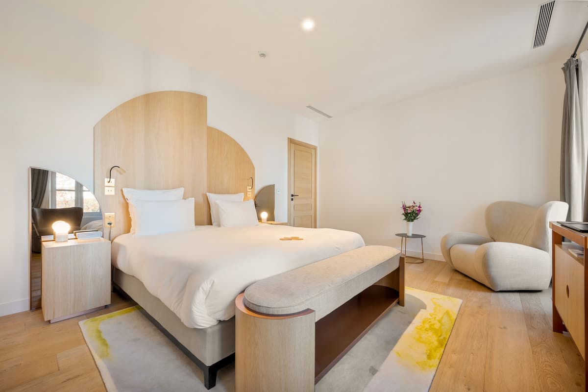 Ein modernes Hotelzimmer mit einem großen Bett, Nachttischen, einer Bank, einem Sessel und einem kleinen Tisch mit Blumen.