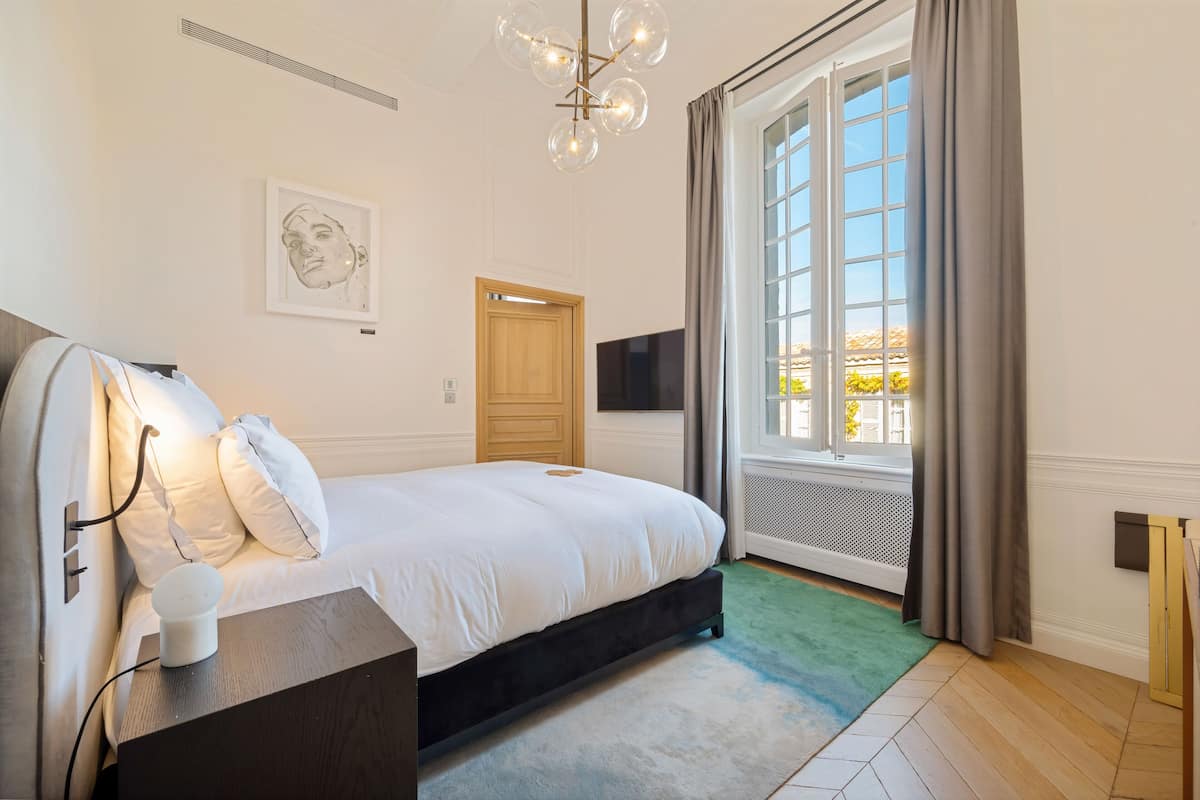 Ein Schlafzimmer mit Bett, Nachttisch, Lampe, Kunstwerk, Fenster und Vorhängen.