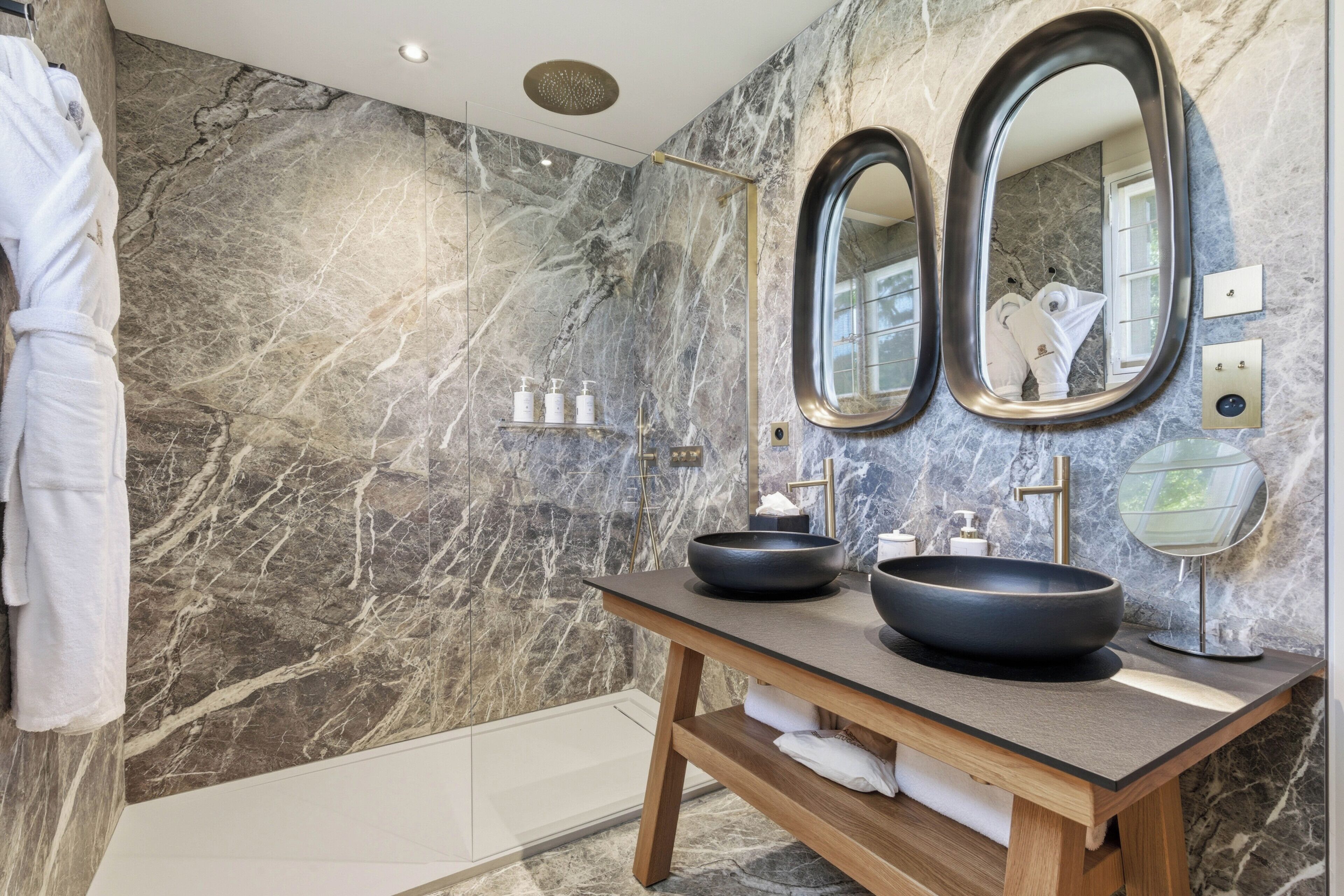 A bathroom with marble walls, a wooden vanity, two large oval mirrors, and a modern sink.