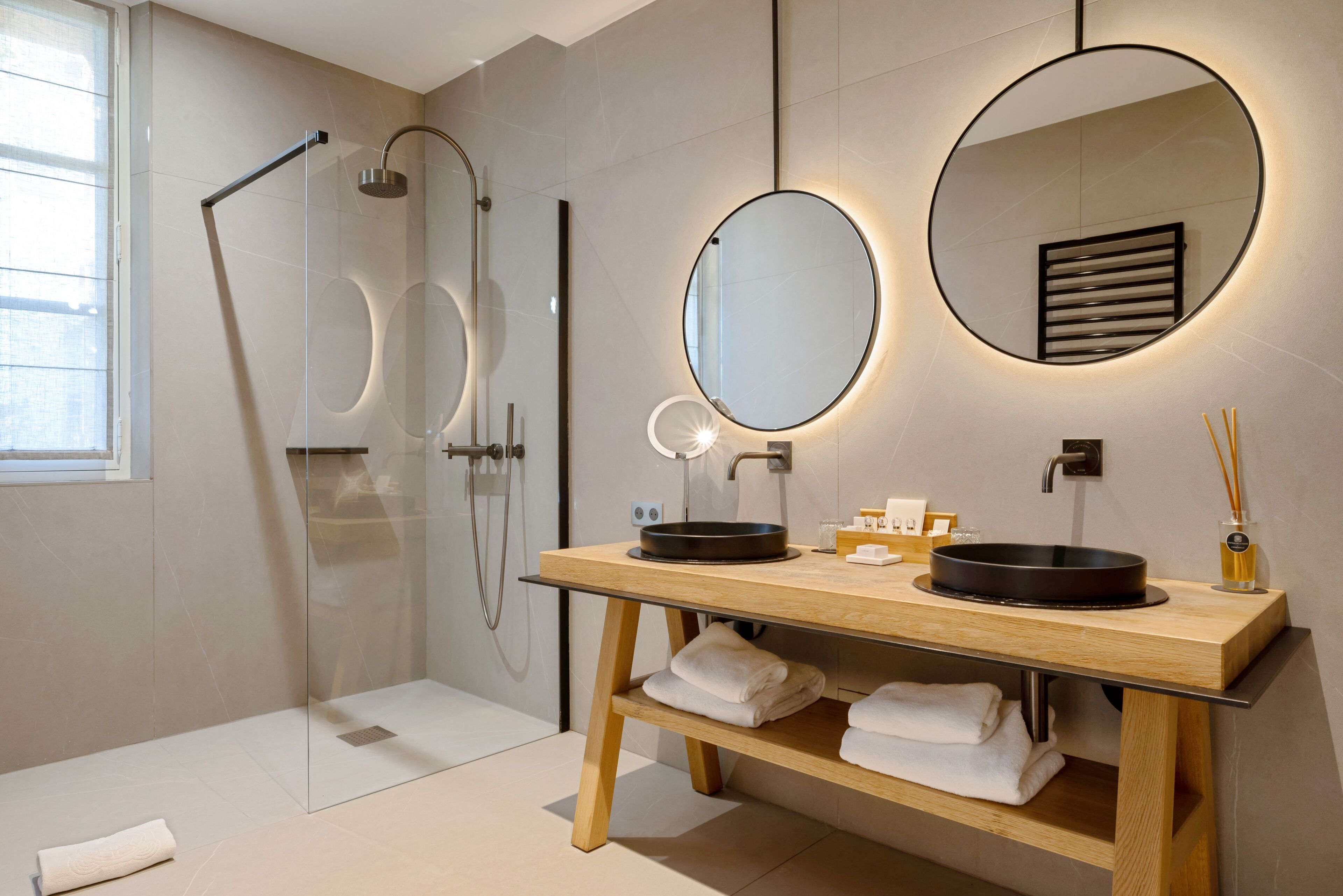 A modern bathroom with a glass-enclosed shower, a wooden vanity with two black sinks, and a round mirror with integrated lighting.