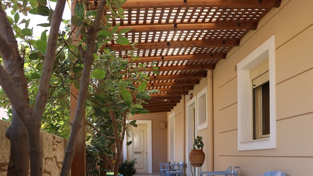 Un couloir extérieur abrité, agrémenté d’une pergola en bois, de plantes en pot et de chaises bleues.