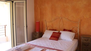 Un lit bien fait, avec du linge de lit blanc et un oreiller rouge, une lampe cylindrique rouge posée sur une table de chevet, et une table de nuit en bois avec des tiroirs.