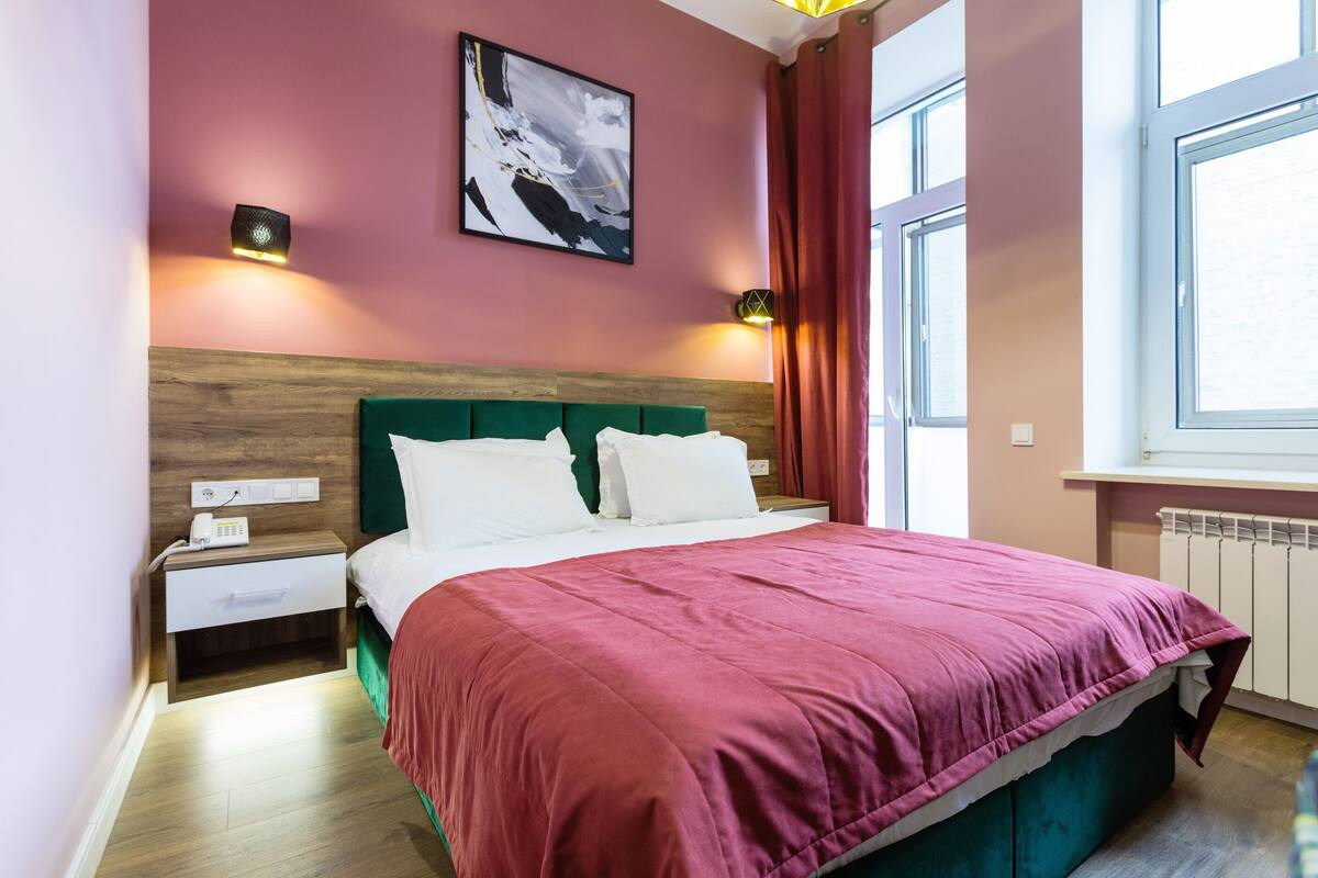 A bedroom with a wooden headboard, a green bedspread, two white pillows, a nightstand with a phone, and a wall-mounted light.