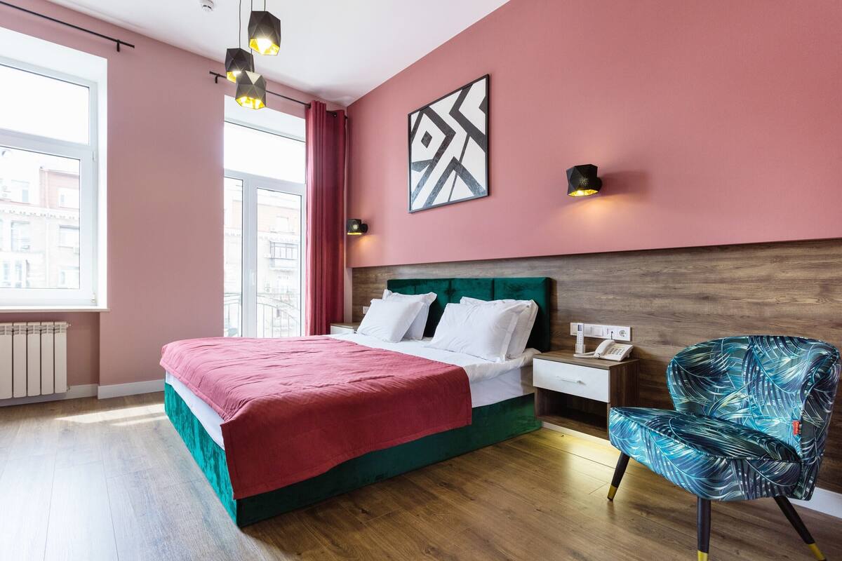 A hotel room with a bed, a chair, a nightstand, a wall art piece, and a window with curtains.