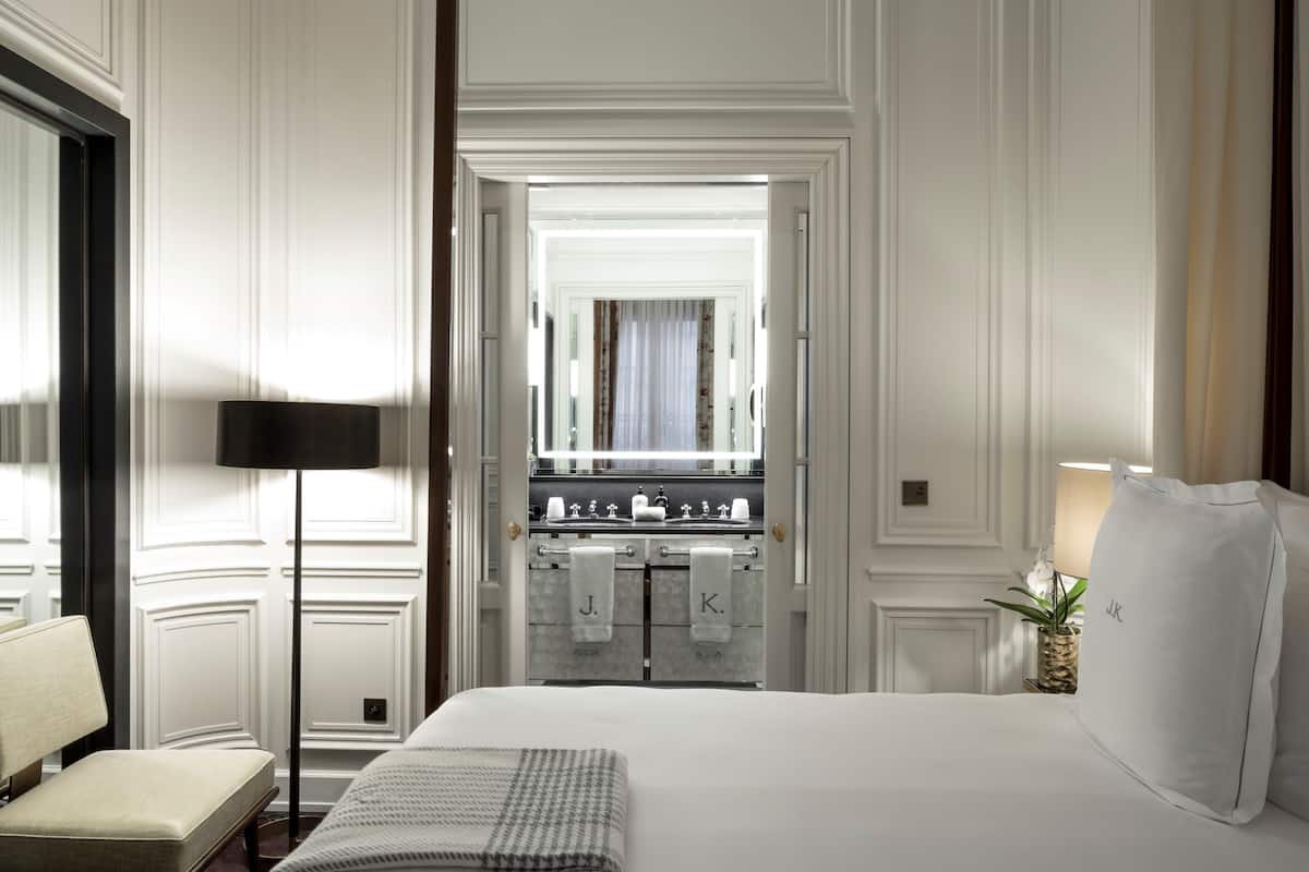 A hotel room with a large bed, a bedside table, a lamp, and a view of the bathroom through an open door.