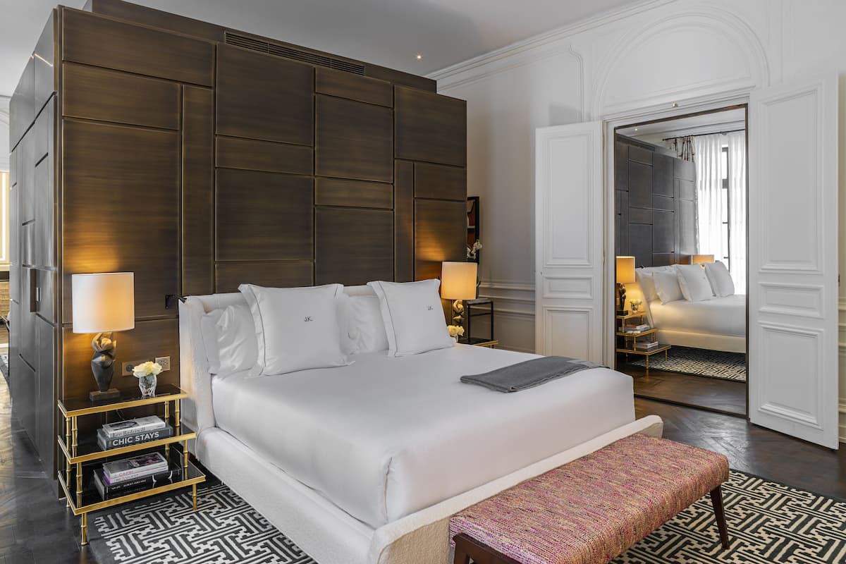 A modern hotel room with a large bed, bedside tables, a bench, and a patterned rug.