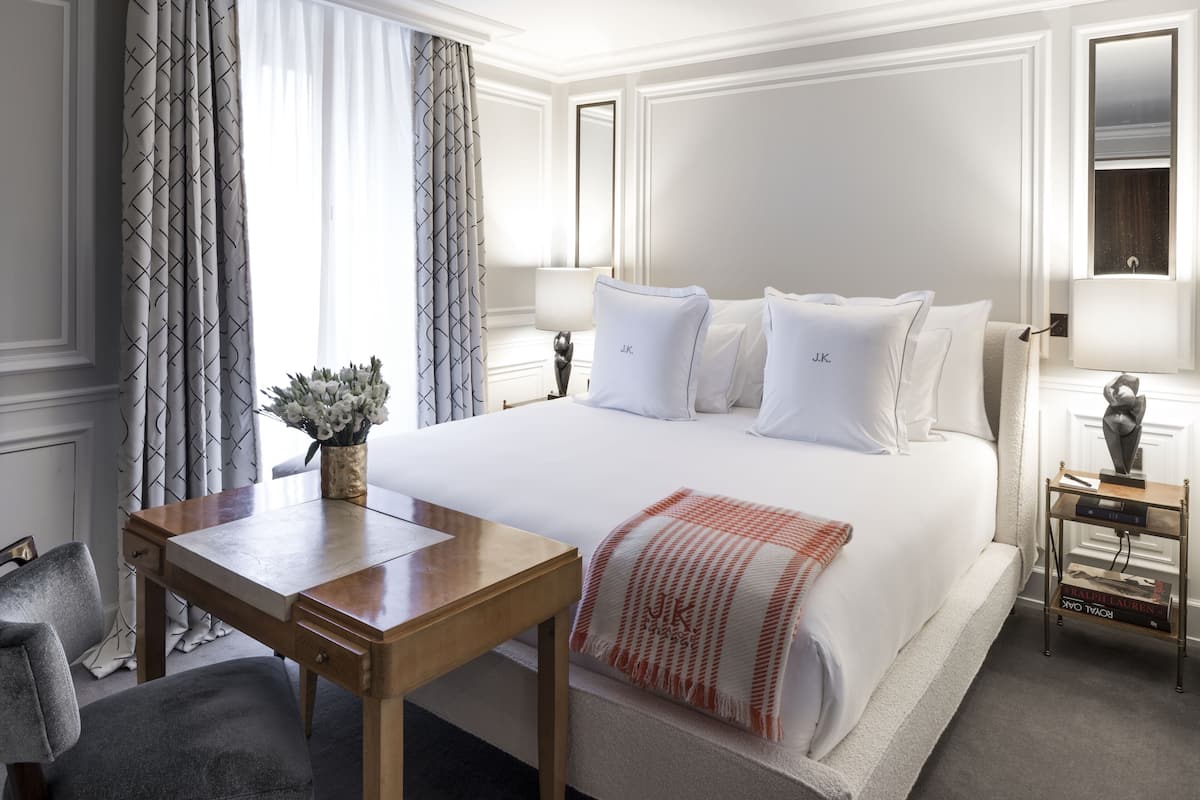 A hotel room with a large bed, a wooden desk, a chair, a nightstand with a lamp, and a mirror.