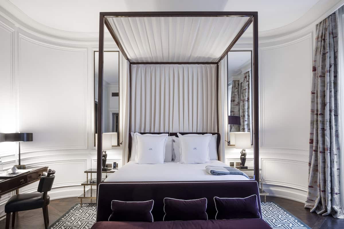 A four-poster bed with white linens and two pillows, a desk with a lamp, and a chair in a hotel room with a patterned rug.