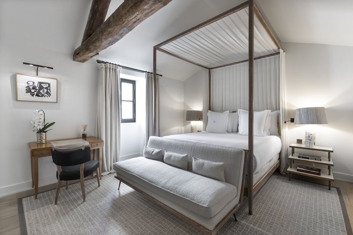 A four-poster canopy bed with a bench in front, a desk with a chair, a small bedside table, and a framed picture on the wall.