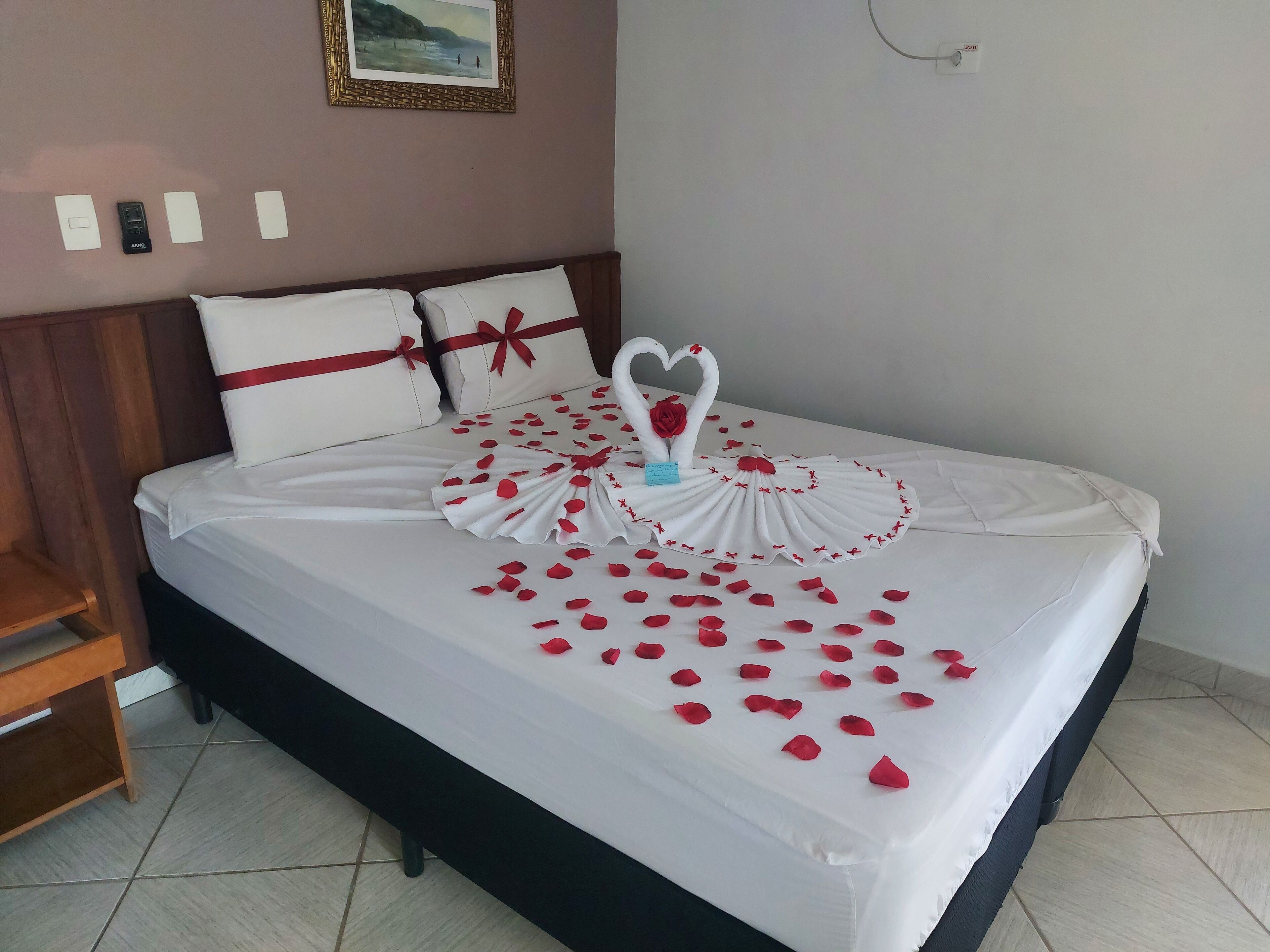 A neatly made bed with a white bedspread, red rose petals, and a heart-shaped decoration made of roses.