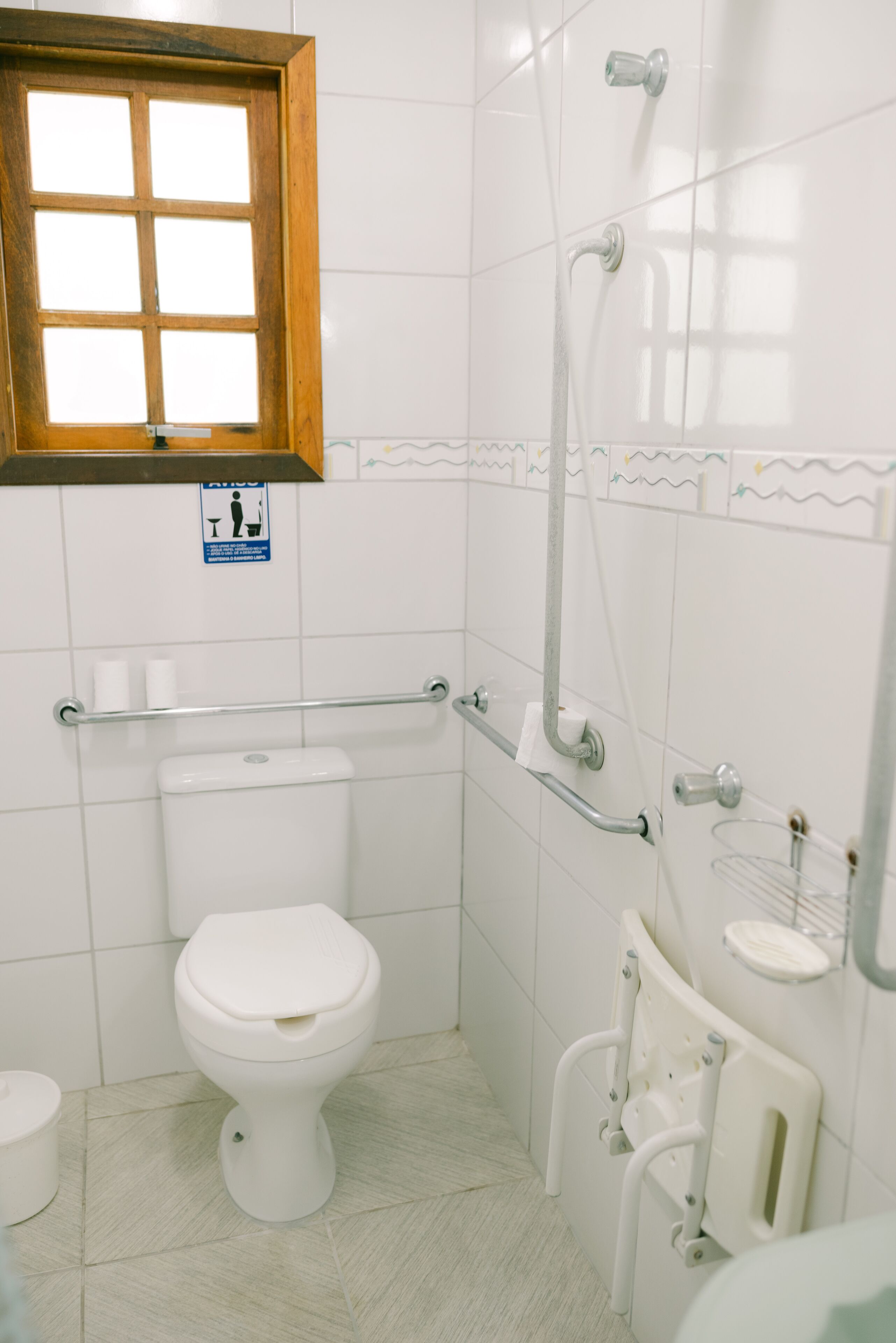 A small, white bathroom with a toilet, grab bars, a shower, and a window.
