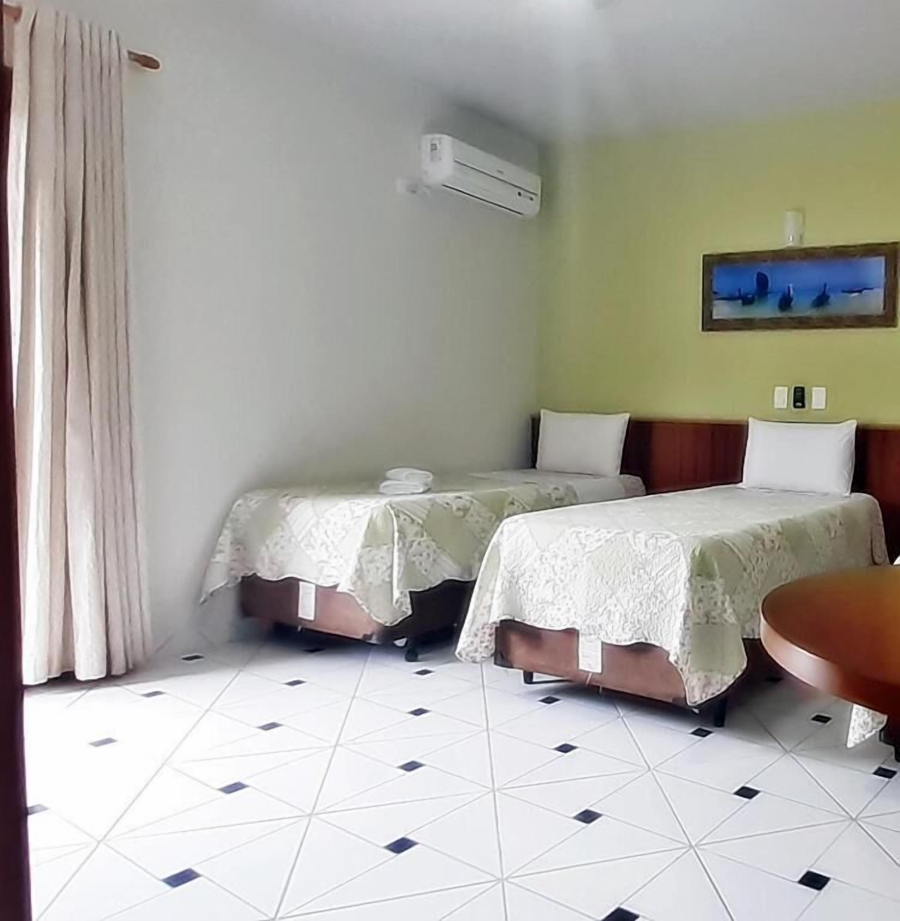 A hotel room with two beds, a table, an air conditioning unit, and a painting on the wall.