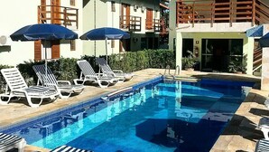 A swimming pool with lounge chairs and umbrellas, surrounded by a building with a red roof and beige walls. - Hotel Kiribati (São Sebastião)