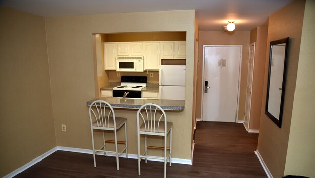 A compact kitchen with white cabinets, a microwave, a stove, and a refrigerator. There are two white chairs at the kitchen counter. - Swan Motel (Linden)