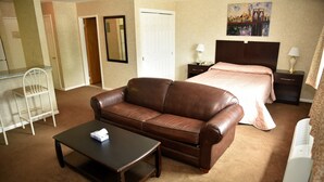 A hotel room with a brown leather sofa, a small table with a tissue box, a bed with a pink quilt, a nightstand with a lamp, and a painting of a bridge on the wall. - Swan Motel (Linden)