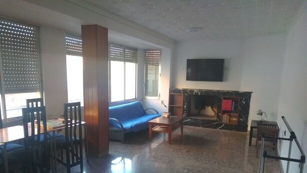A living room with a sofa, a fireplace, a television mounted on the wall, and a dining area with chairs and a table. - Habitaciones Petrer (Petrer)