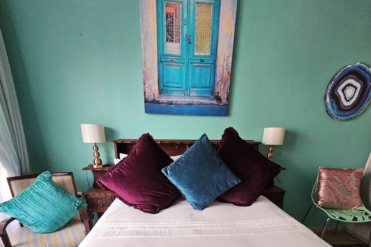 Una cama con edredón blanco, dos almohadas, una manta verde y un cuadro de una puerta azul en la pared.