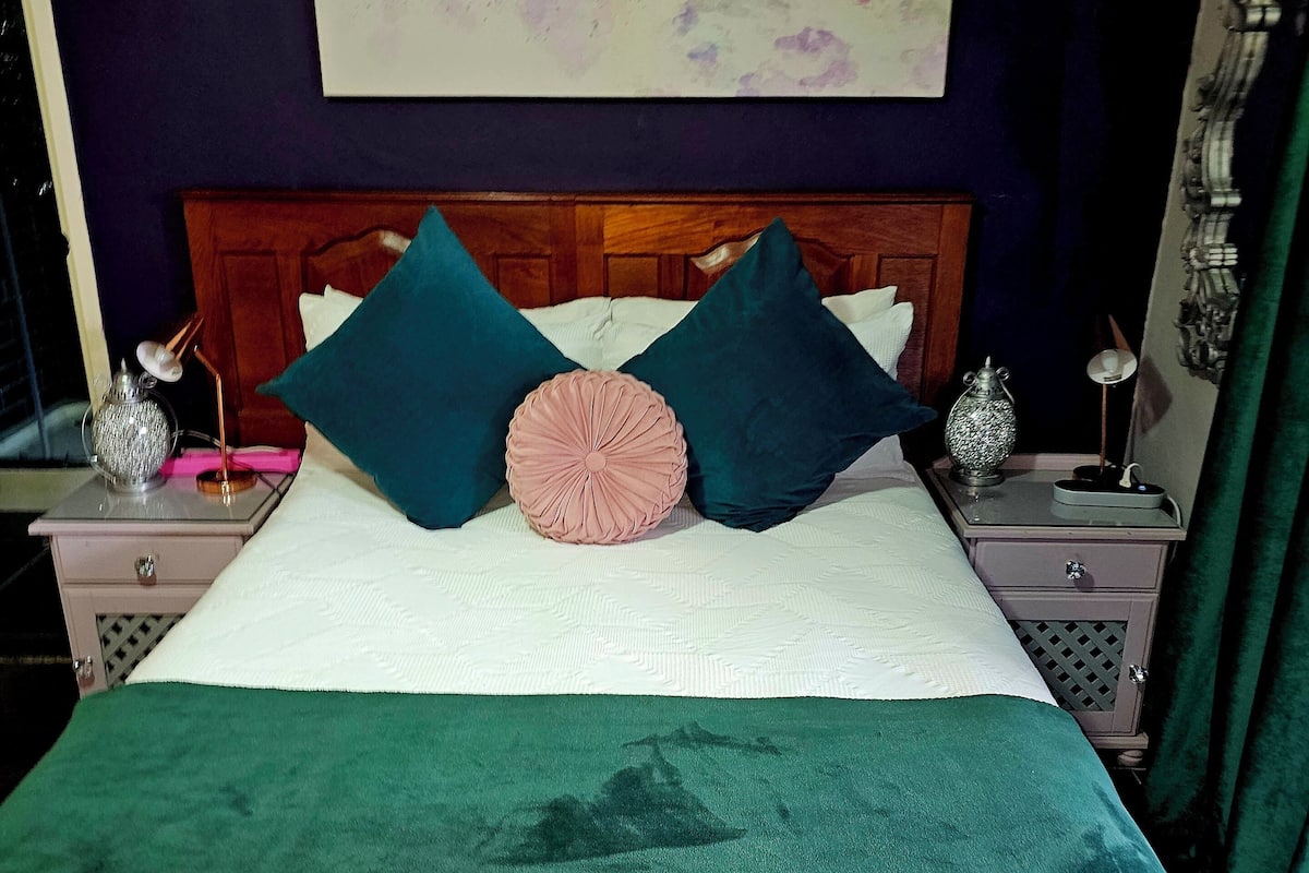 Una cama bien hecha con una manta verde, dos almohadas color turquesa y una almohada decorativa rosa. Se observa un cabecero de madera, una mesita de noche con una lámpara y un cuadro colgado en la pared.