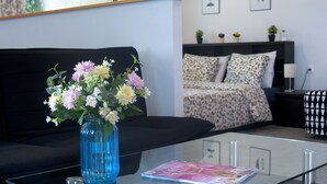 A modern living room with a glass coffee table, a blue vase with flowers, a sofa, a bed with patterned bedding, and a bedside table with a lamp. - Cozy Fully Renovated Studio in Holargos (Papagou-Holargos)