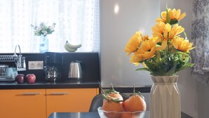 A kitchen with a fruit bowl containing oranges and apples, a vase with yellow flowers, and a window with curtains. - Cozy Fully Renovated Studio in Holargos (Papagou-Holargos)