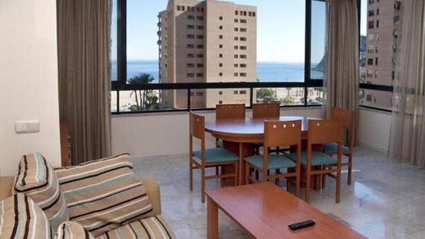 A hotel room with a large window, a dining table with chairs, a sofa, and a remote control on the table. - Paraiso Florida (Benidorm)