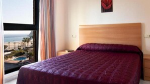 A bedroom with a large bed, a wooden headboard, a nightstand, a painting, and a view of the beach and pool through a window. - Paraiso Florida (Benidorm)