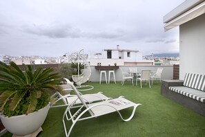 A rooftop terrace with artificial grass, white lounge chairs, a striped sofa, and a small table with chairs.