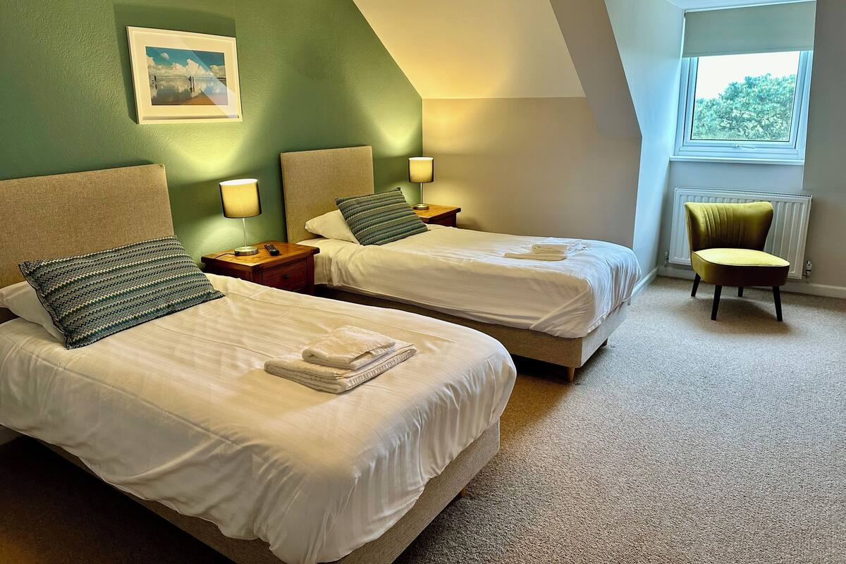 Quarto de hotel com duas camas, parede verde, quadro emoldurado e janela com vista para árvores.
