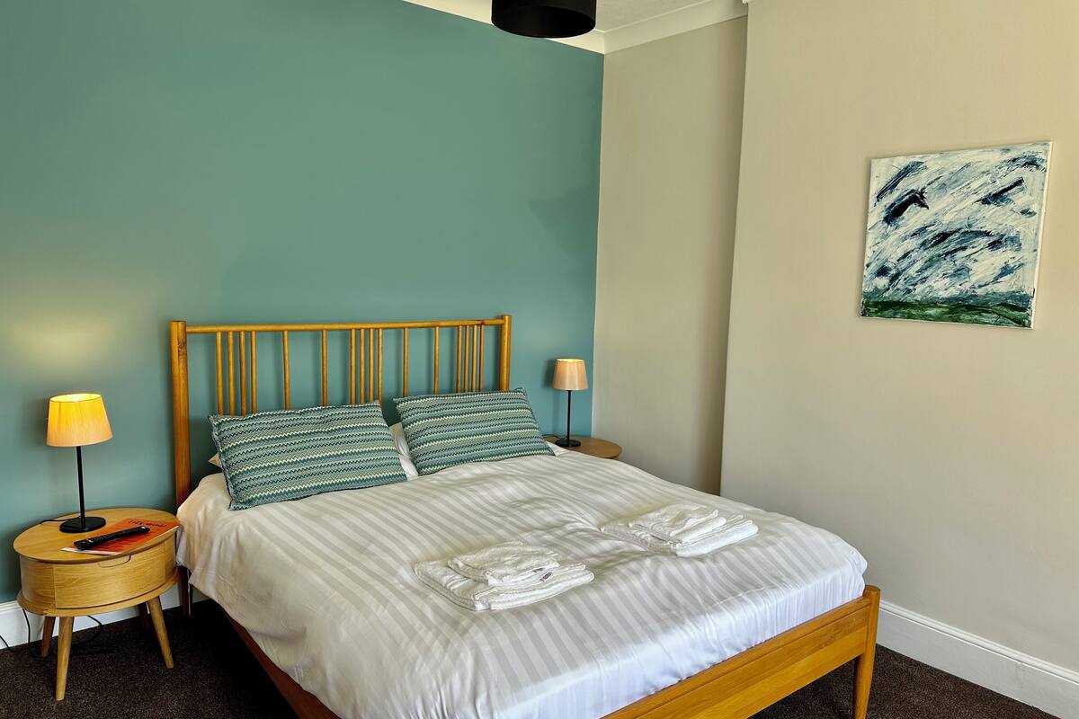 Uma cama bem arrumada com roupa de cama listrada e duas almofadas, um abajur e um controle remoto em uma mesa de cabeceira de madeira.
