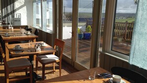 A dining area with wooden tables and chairs, large windows with a view of solar panels and a fence. - St George's Hotel (Perranporth)