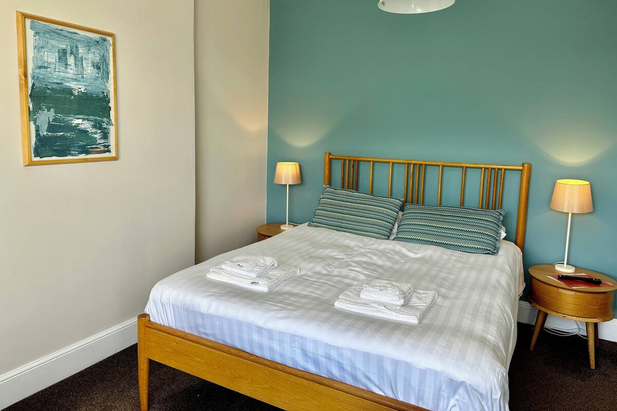 Cama bem arrumada com lençóis brancos e duas almofadas, cabeceira de madeira, abajures de cabeceira e uma obra de arte emoldurada na parede.