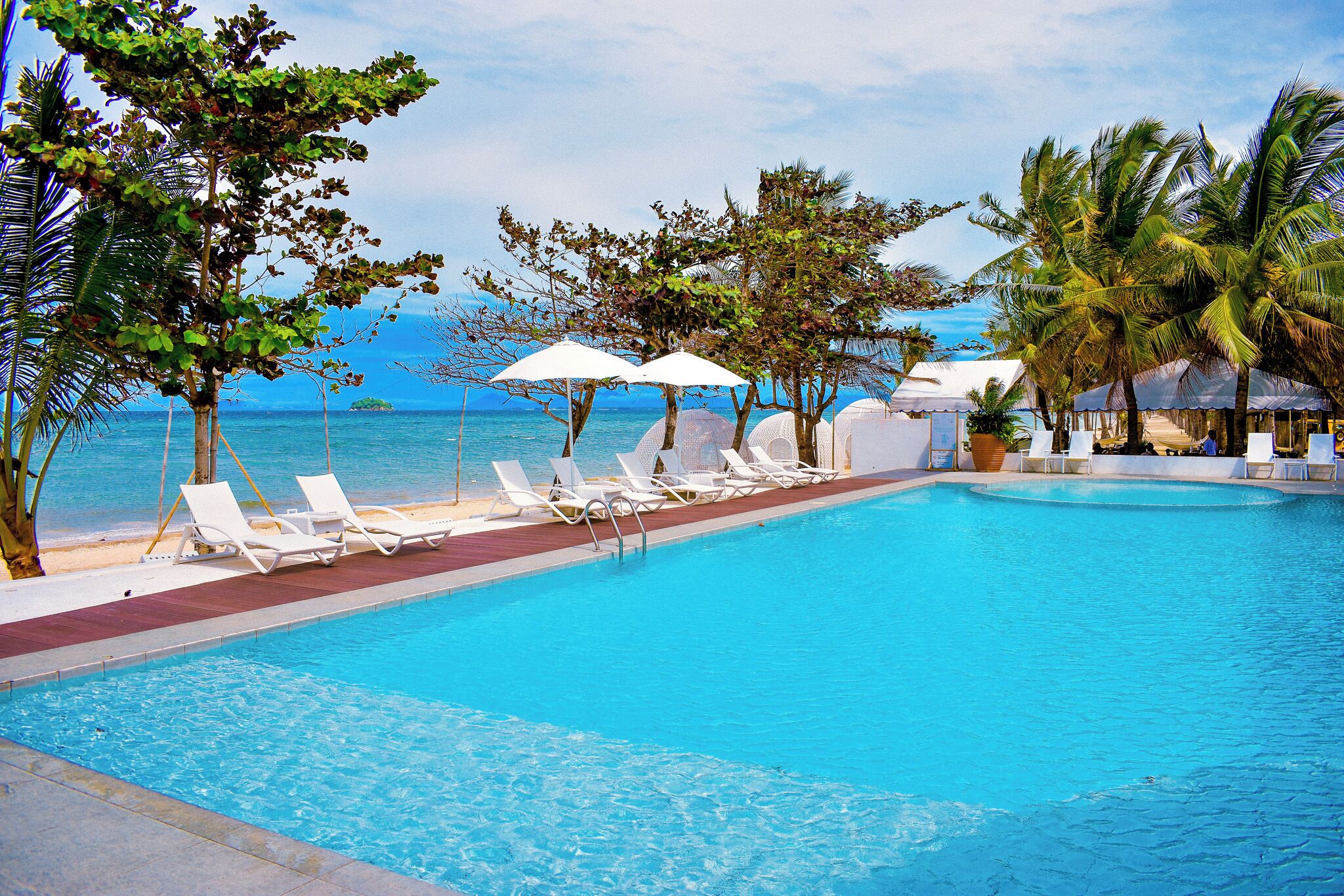 A pool with lounge chairs and umbrellas, surrounded by palm trees and a view of the ocean.