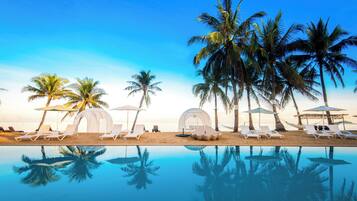 Une piscine située au bord de la plage, avec des chaises longues, des parasols et des palmiers.