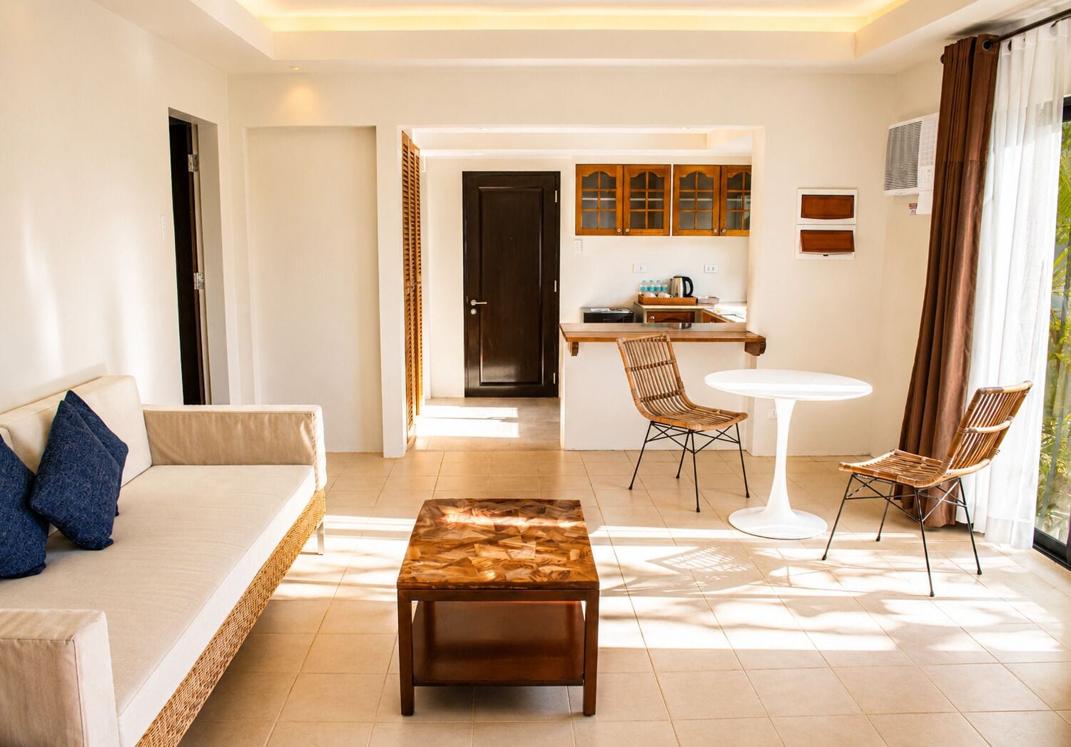 A modern hotel room with a sofa, a coffee table, two wicker chairs, a dining table, and a kitchen area.