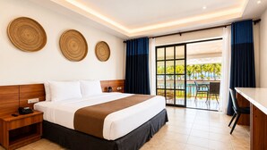 A hotel room with a bed, bedside table, and a desk with a chair. There are large windows with curtains, and a view of palm trees and a pool. - Golden Sands Destination Resorts (Daanbantayan)