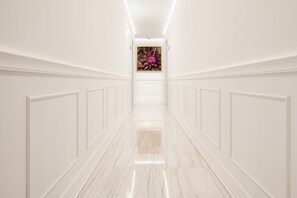 A long, narrow hallway with white walls and a marble floor, leading to a room with a large abstract painting. - Flos Luxury Rooms (Split)