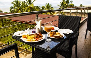 Una mesa de comedor con platos de comida, un vaso de jugo y una botella de agua, ubicada en un balcón con vista a palmeras y al océano.