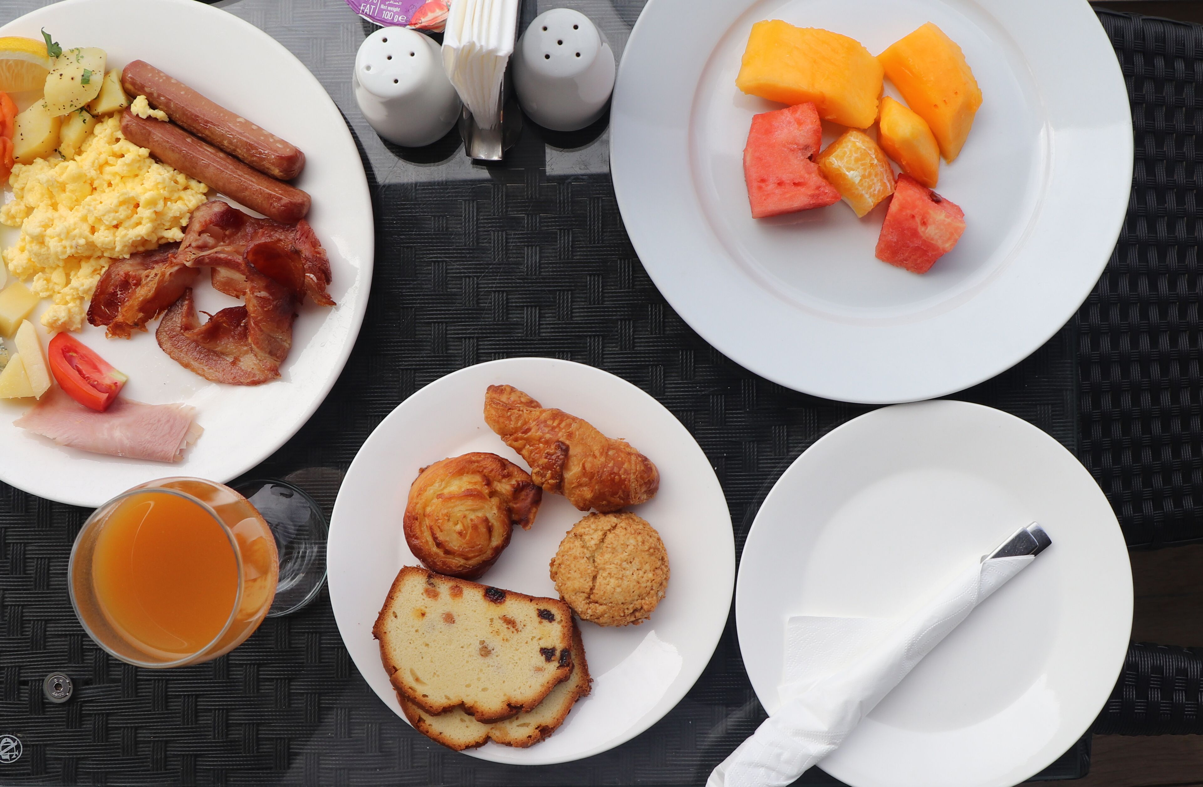 A breakfast setting with plates of eggs, bacon, sausages, and toast, along with a glass of orange juice.