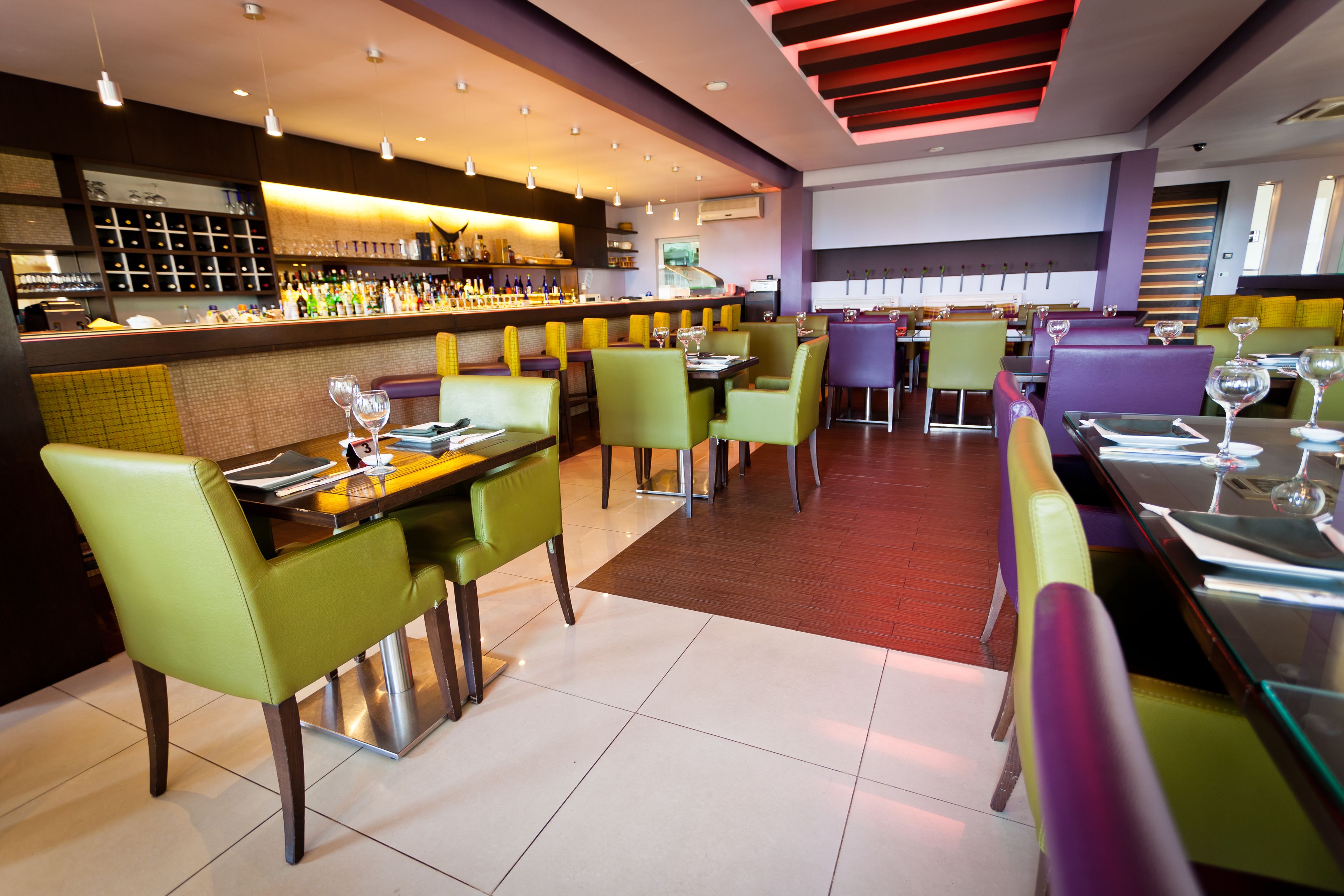 Un interior de restaurante moderno con varias mesas y sillas, una zona de bar con estantes llenos de botellas y un suelo de baldosas impecable.