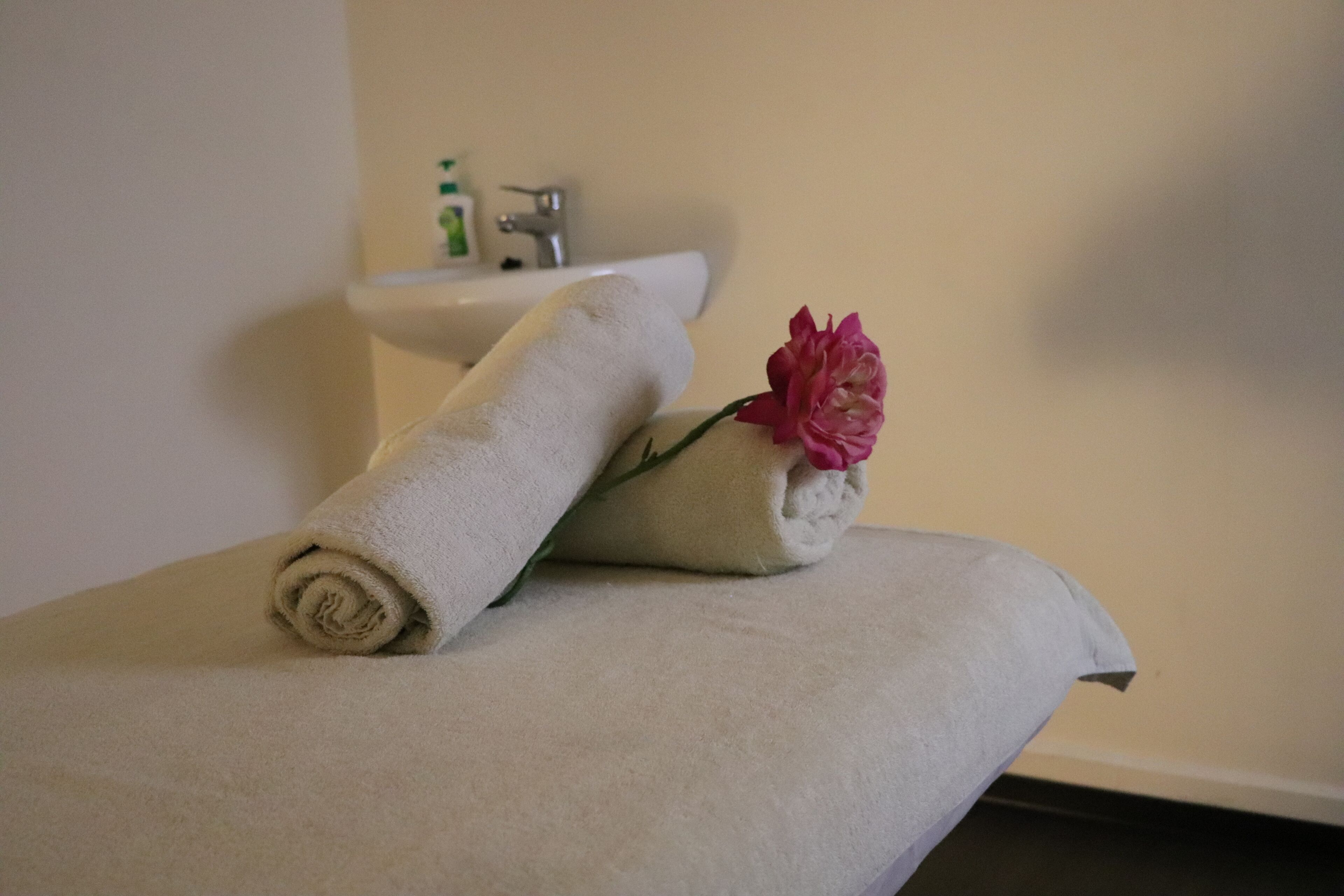 Una toalla color beige doblada con cuidado y una flor rosa sobre una superficie blanca, probablemente un mostrador de baño, con un lavamanos y un estante pequeño al fondo.