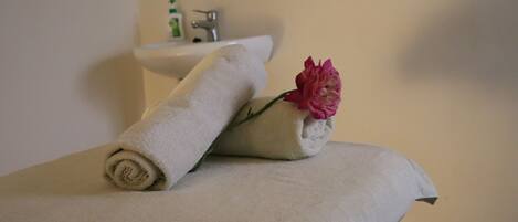 Una toalla color beige doblada con cuidado y una flor rosa sobre una superficie blanca, probablemente un mostrador de baño, con un lavamanos y un estante pequeño al fondo.