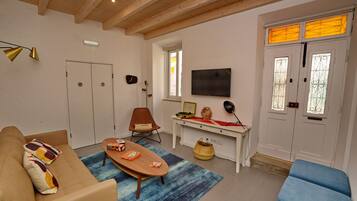 A living room with a sofa, coffee table, and a TV on the wall. There is a desk with a computer and a lamp. A door with a window is visible in the background.
