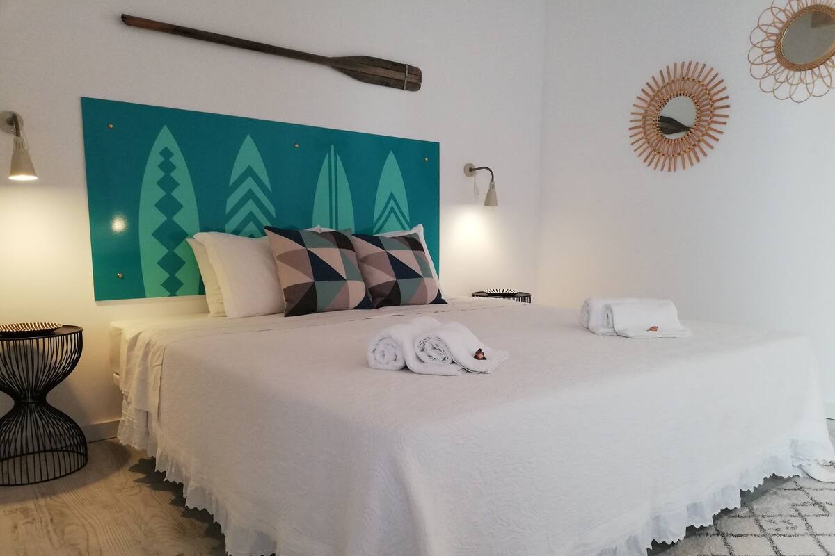 A neatly made bed with white linens and rolled towels, a teal wall with geometric patterns, a wooden headboard, and a round mirror on the wall.